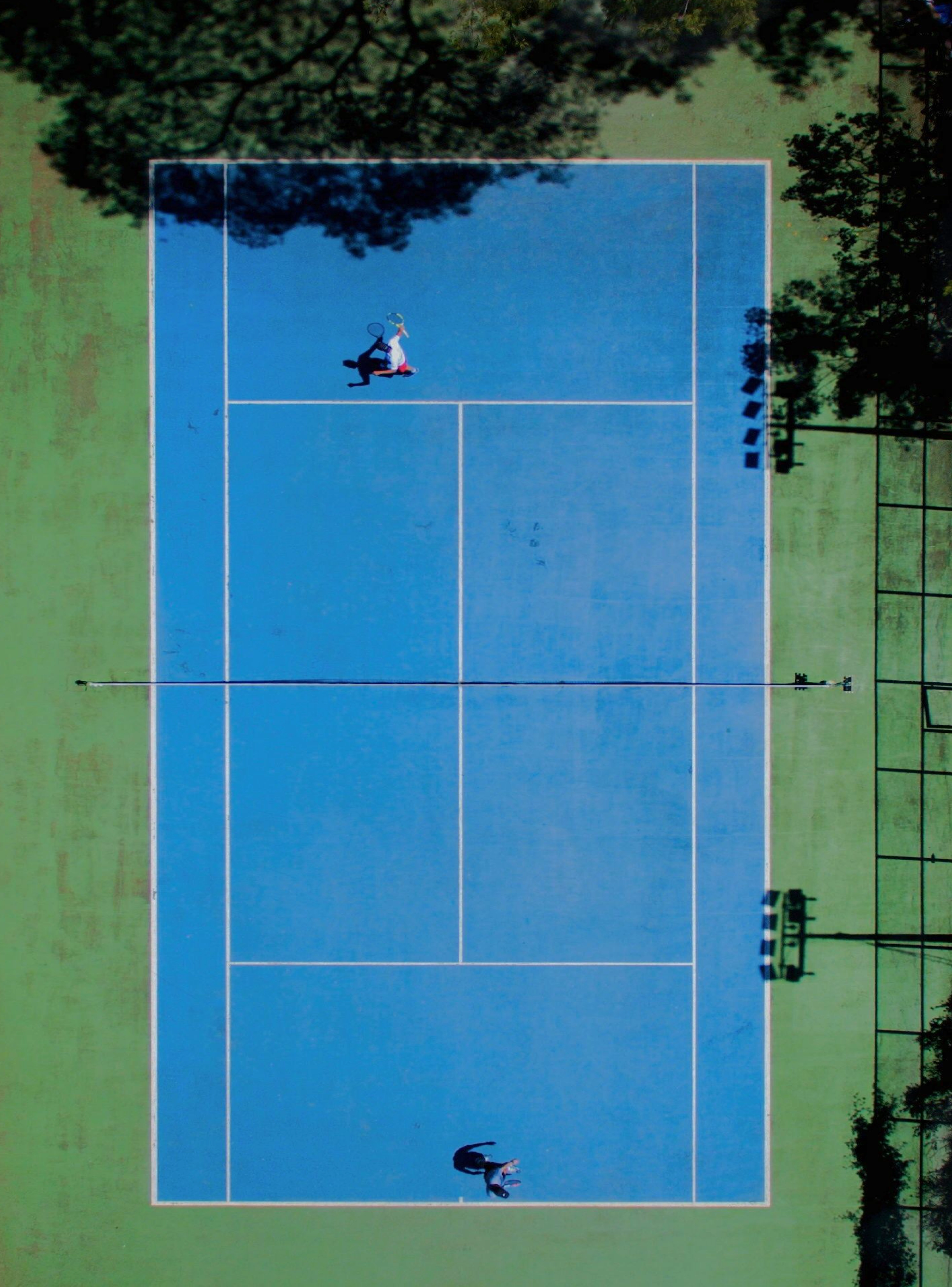 Vista aérea de um campo de ténis azul rodeado por áreas verdes, com dois jogadores em lados opostos.