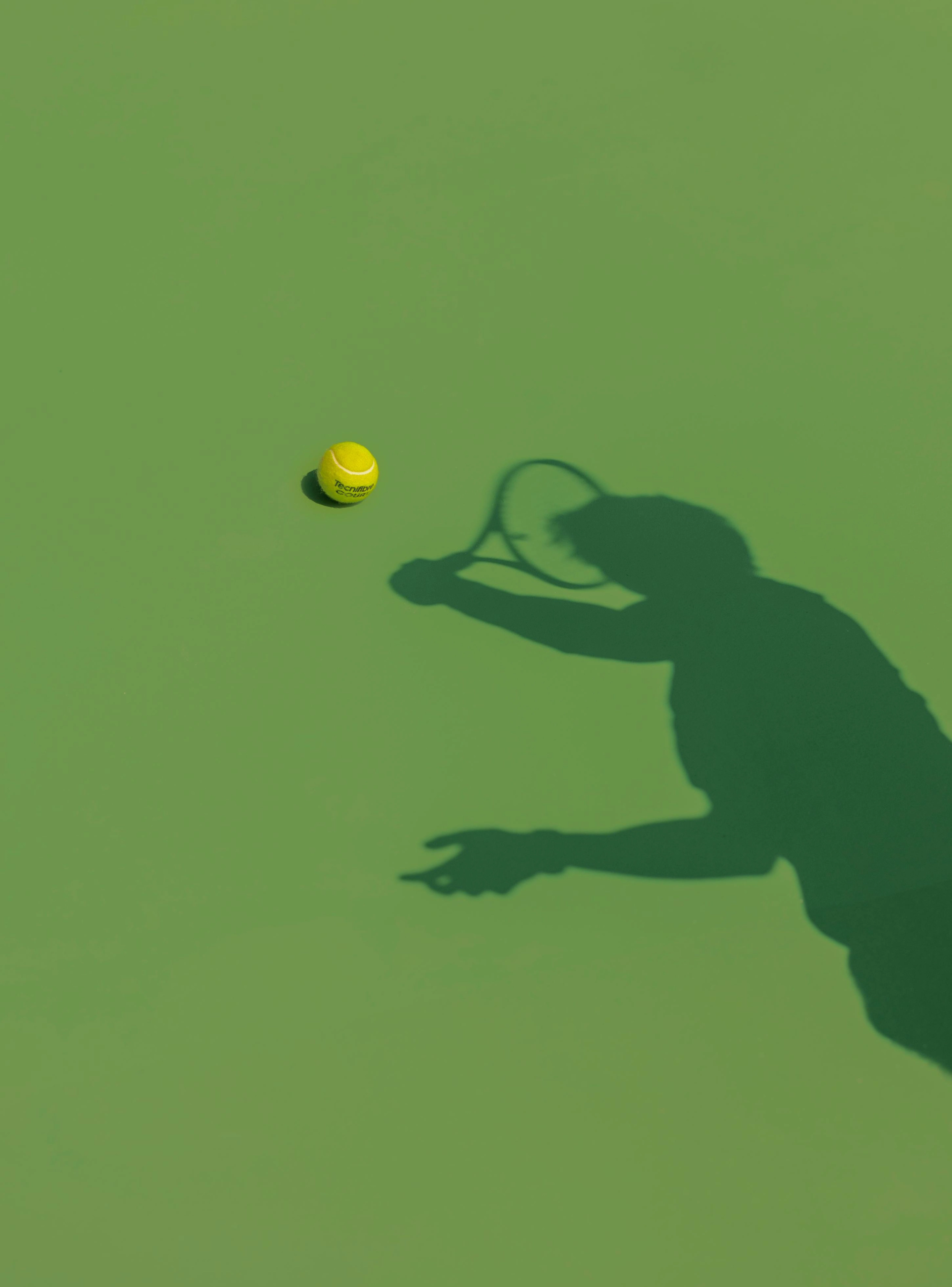 Sombra de um jogador de ténis a preparar-se para bater a bola, com a bola amarela em destaque num campo verde.
