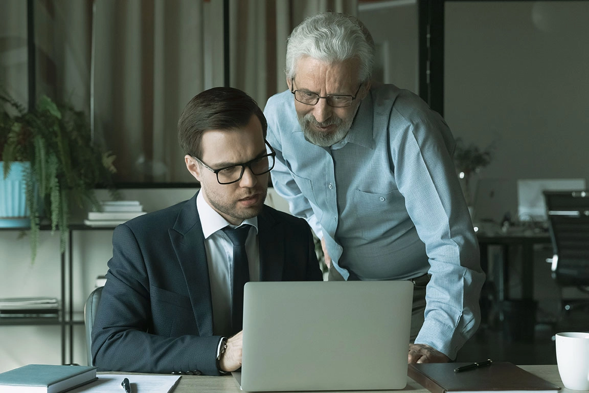 Pessoa mais jovem no computador a falar com outra mais velha ao lado, ambos vestidos formalmente em ambiente de trabalho, simbolizando os serviços experientes de Cobertura de Risco