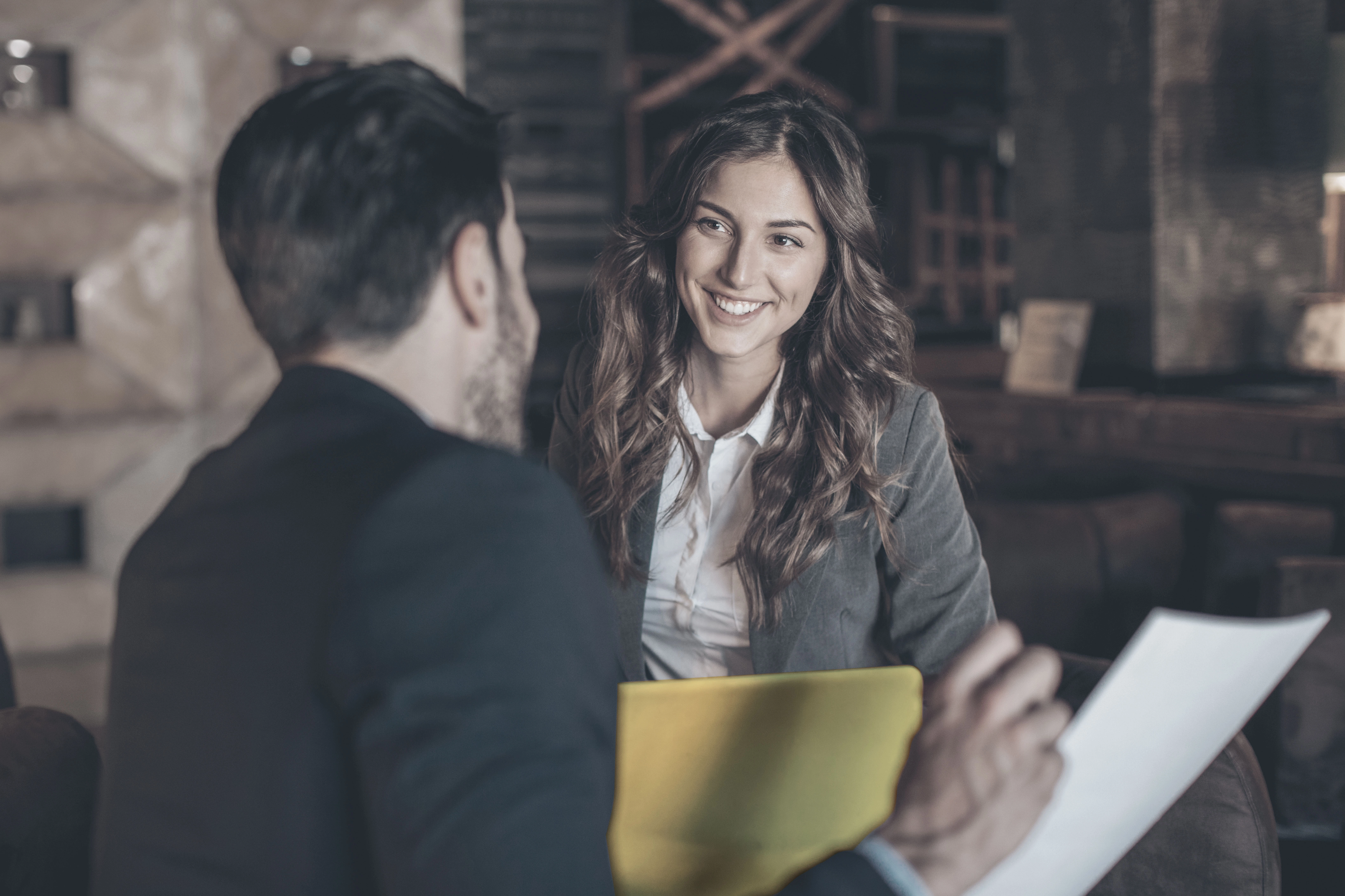 Conversa entre duas pessoas vestidas formalmente, com foco numa mulher que se encontra feliz, simbolizando o desenvolvimento de opções que beneficiem o cliente do Banco Carregosa na obtenção dos melhores resultados