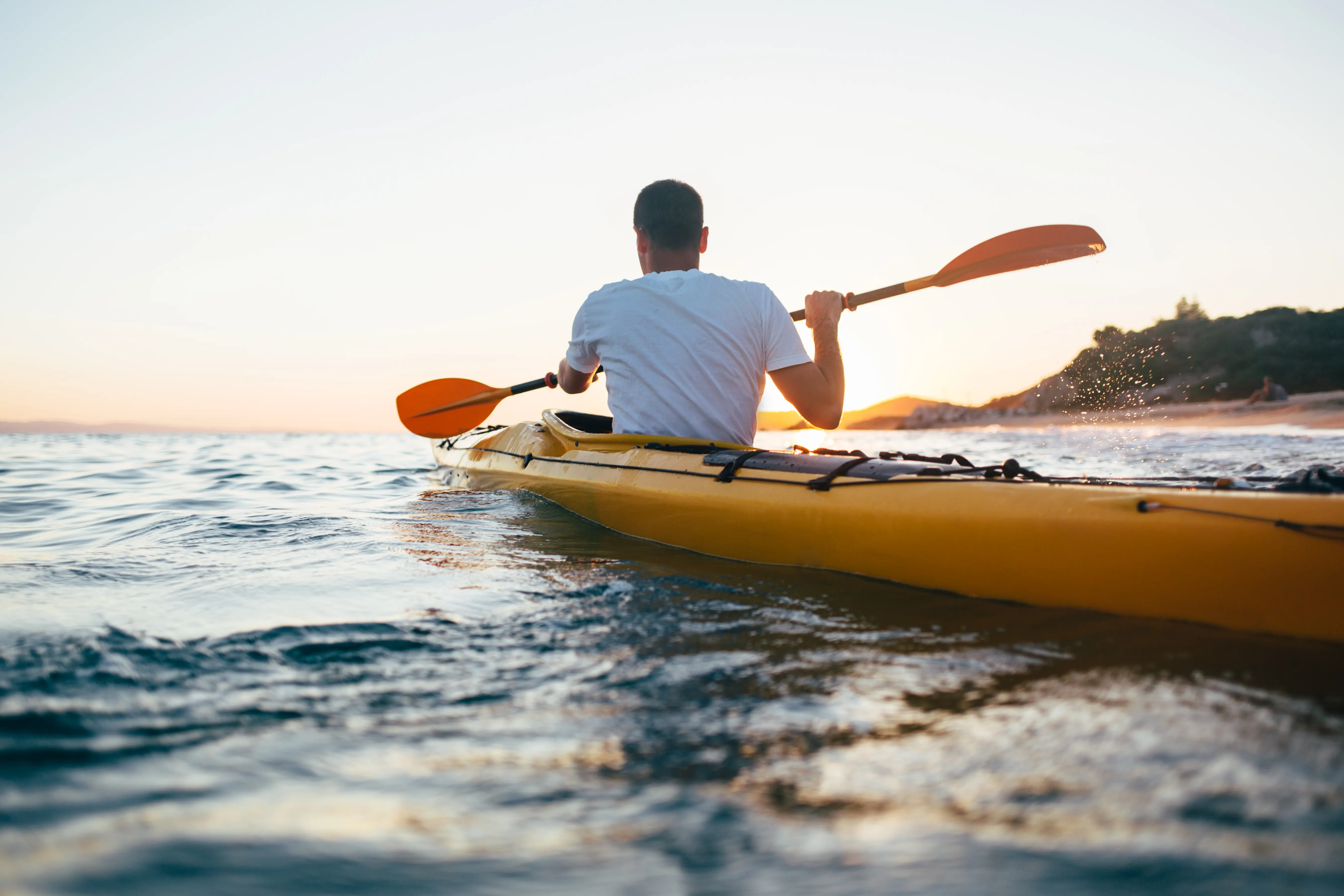 Um homem visto de costas, a remar num caiaque amarelo no oceano ao pôr do sol.