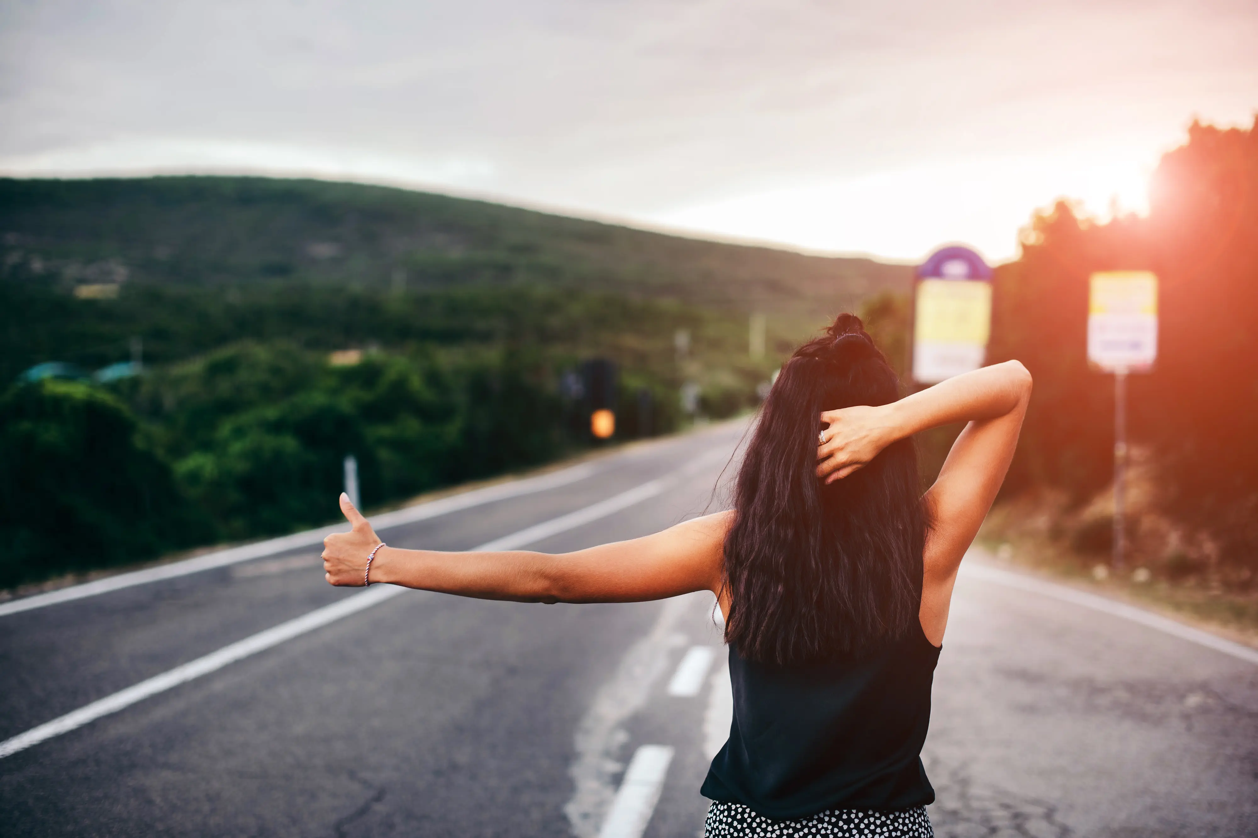 Mulher de costas a fazer autostop numa estrada ao pôr do sol.
