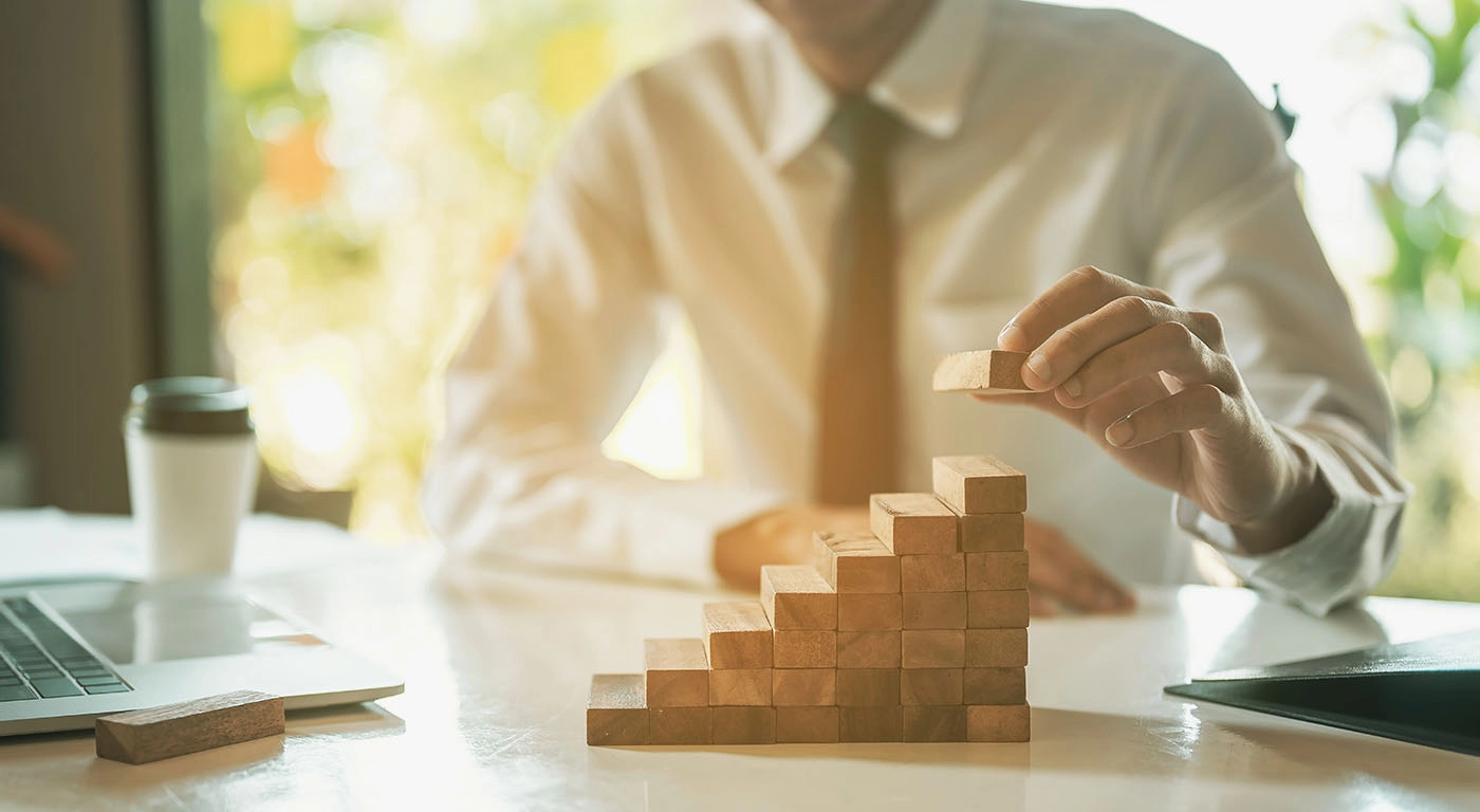 Homem vestido formalmente, em contexto de trabalho, a colocar blocos de madeira uns em cima dos outros criando um crescimento gradual de escadas