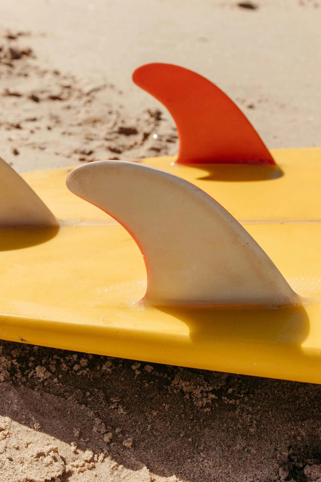 Plano aproximado das quilhas cor de laranja de uma prancha de surf amarela, pousada na areia molhada, simbolizando a fiabilidade de investir em ETFs.