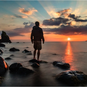 Homem de pé sobre rochas na água, olhando para o pôr do sol sobre o oceano, com raios de luz a emanar das nuvens.