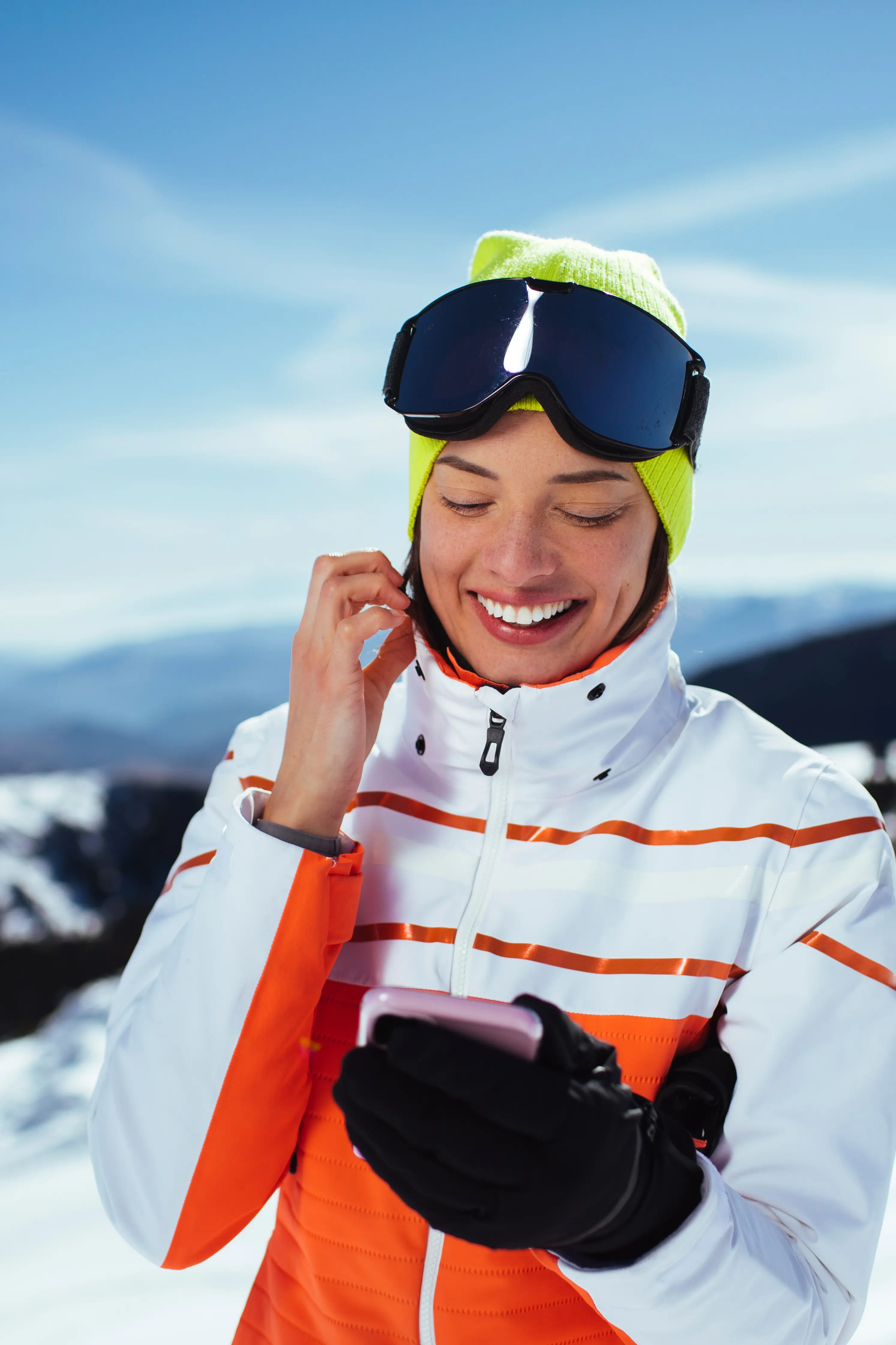 Mulher sorridente a usar um telemóvel numa estância de esqui, vestida com um casaco de inverno branco e laranja, gorro amarelo e óculos de esqui pousados na testa, com montanhas cobertas de neve ao fundo.