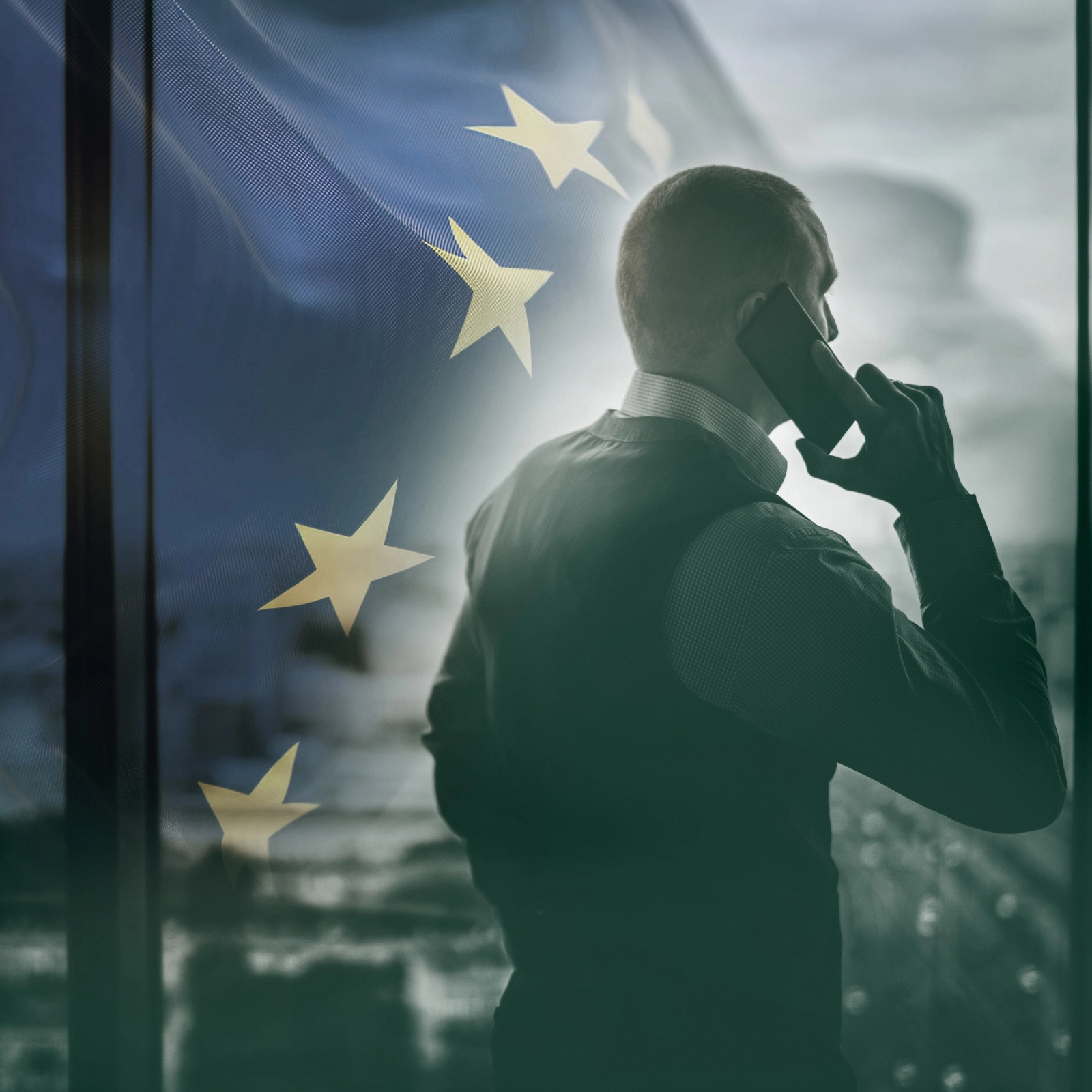 Homem de costas, vestido formalmente, a falar ao telemóvel e a olhar para a paisagem do escritório, com sobreposição da bandeira da união europeia