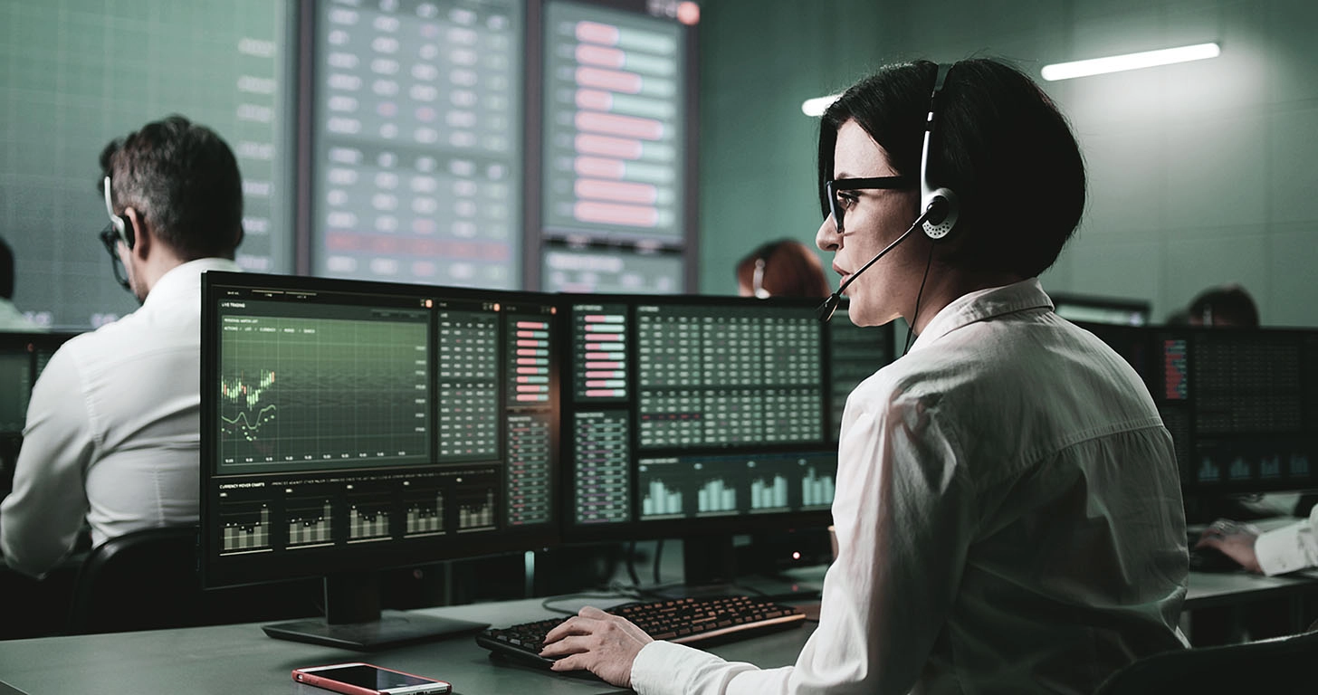 Woman in a workplace with multiple people, wearing headphones and analyzing graphs on two monitors, symbolizing the analysis and management of Structured Products for portfolio diversification and capital protection.