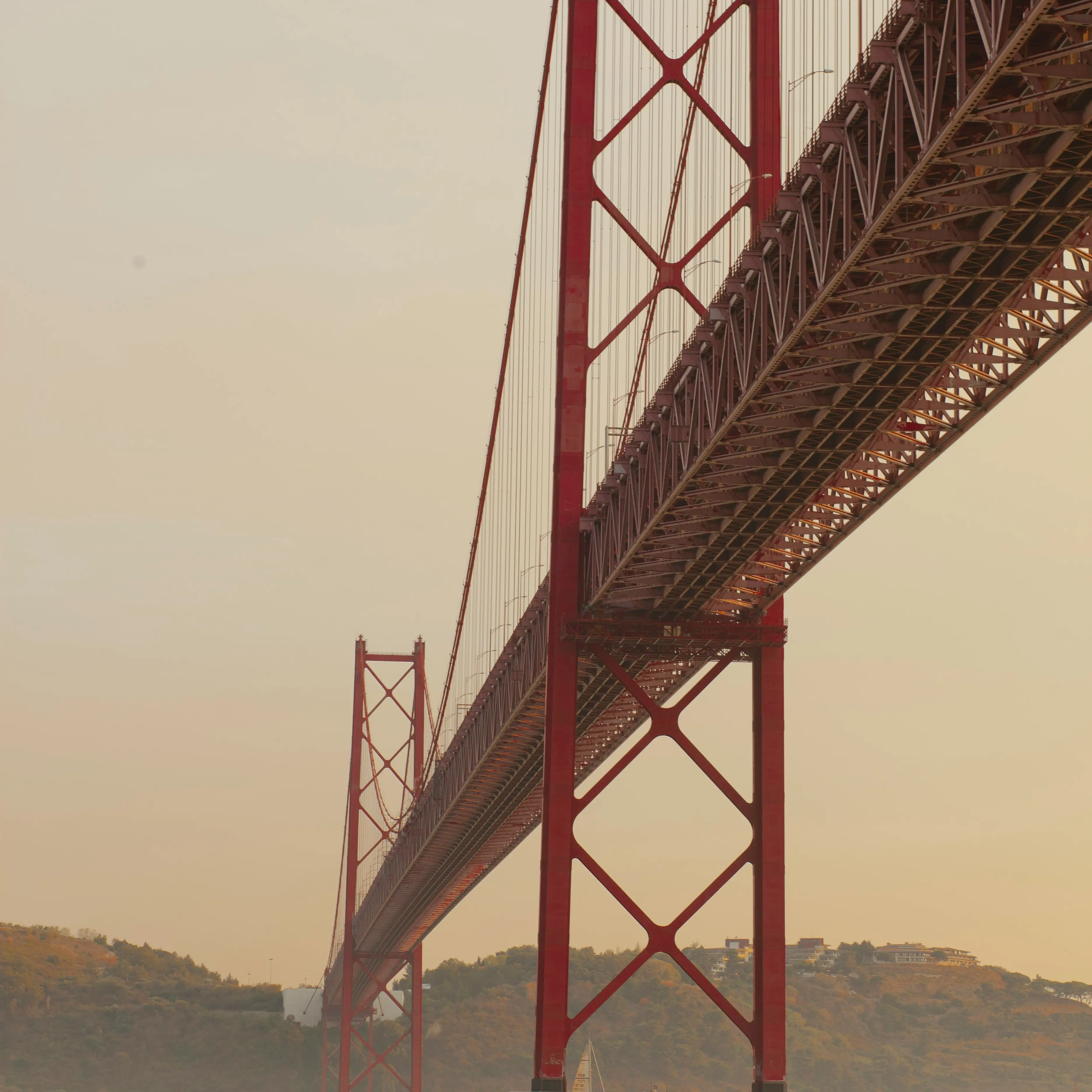 Red suspension bridge viewed from below, spanning a wide river, with tall steel towers and cables, softly lit by late-afternoon light, hills in the background, and a small sailboat on the water.