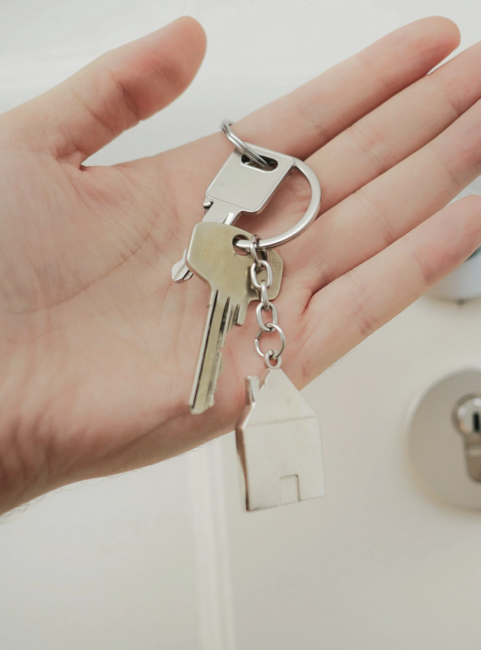Mão a segurar um porta-chaves com duas chaves de metal e um pequeno amuleto em forma de casa, simbolizando a compra de casa.