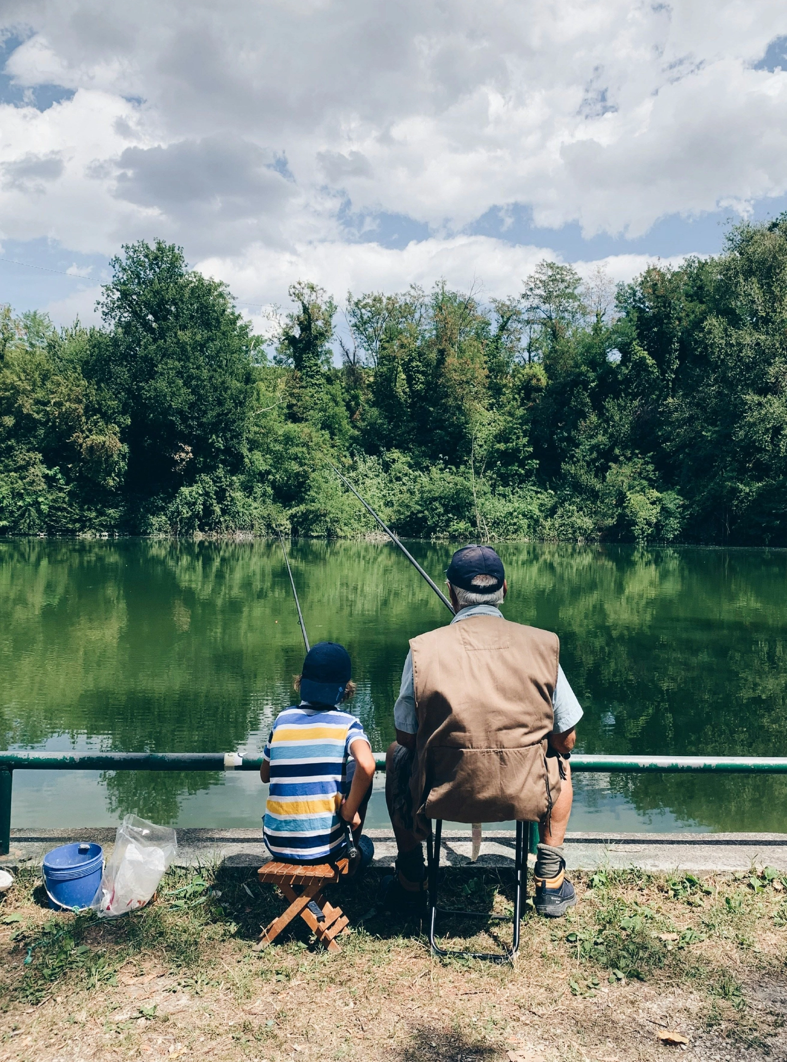 Um homem e uma criança sentados num banco junto a um lago sereno, rodeado de relva e árvores exuberantes.